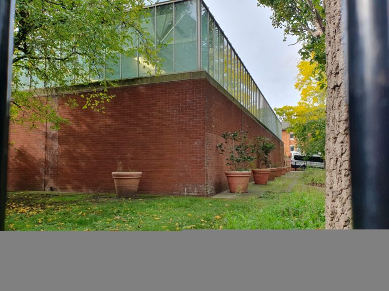 View through a metal fence of a brick wall with a glass railing above; potted plants line the walkway and trees surround the scene.