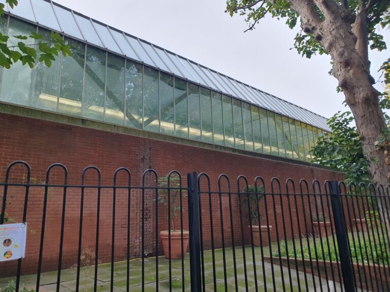 Brick wall with a long glass-roofed addition and metal frame, viewed through a black fence; a large tree trunk on the right.