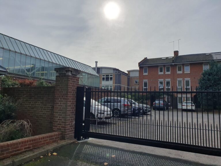 Brick apartment building beside a glass-paneled conservatory, viewed over a black metal gate with parked cars in the foreground.