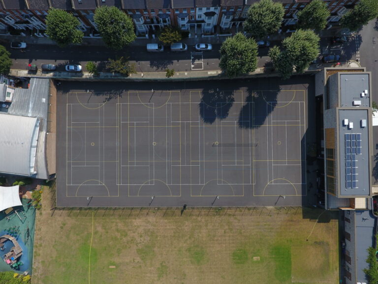 Aerial view of a large outdoor sports court complex with multiple yellow and white boundary lines, adjacent to a road with parked cars and row houses along the street.