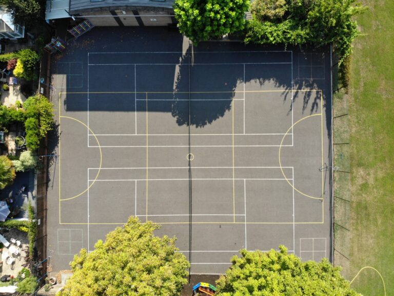Aerial view of an outdoor basketball court with white and yellow boundary lines, surrounded by trees and buildings.