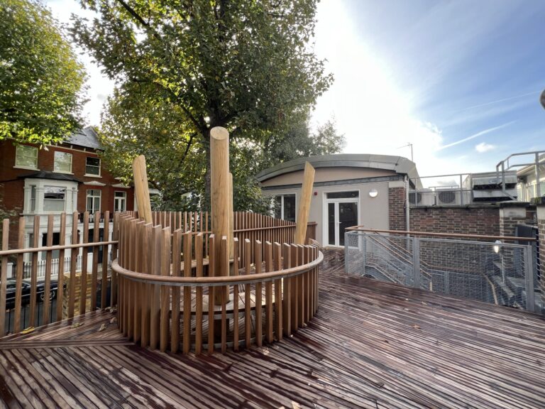 Circular wooden deck with vertical slats surrounding a central post on an urban rooftop playground with brick houses and trees in the background, blue sky above