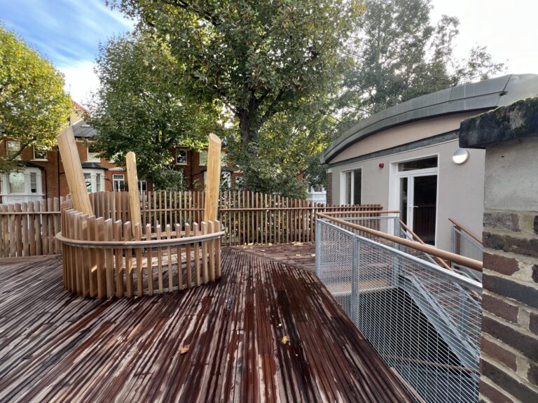 Curved wooden deck and circular bench area surrounded by a tall wooden fence, with trees and brick houses in the background.