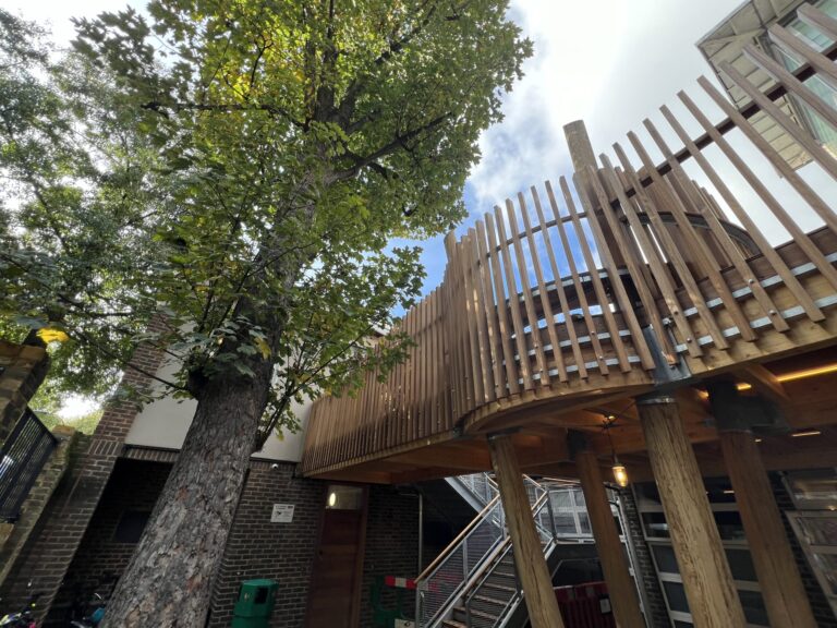 Low-angle view of a brick building courtyard with a tall wooden slatted railing and a large tree overhead against a blue sky.