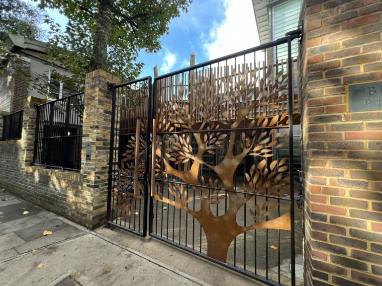Rusty metal gate with a tree-branch silhouette between brick pillars along a sidewalk, blue sky above.