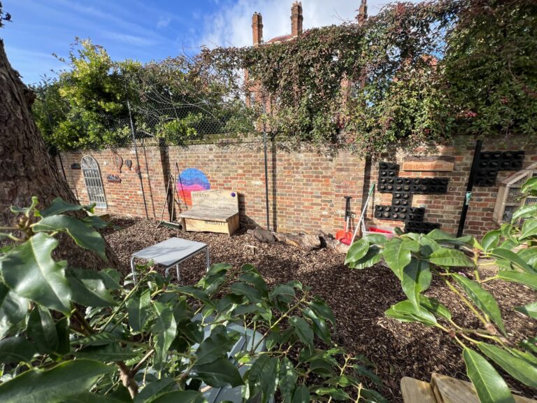 Brick courtyard with ivy-covered wall, chain-link fence, and a wooden bench against a colorful mural.