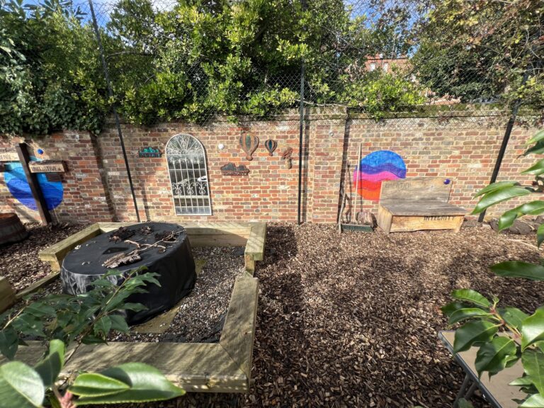 Outdoor garden scene with brick wall, chain-link fence, dense green foliage, and wooden benches.