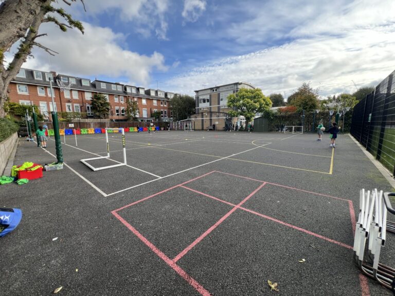 School playground with kids playing on a multi-sport court; pink court lines, colorful seating barriers, and surrounding brick buildings under a cloudy sky