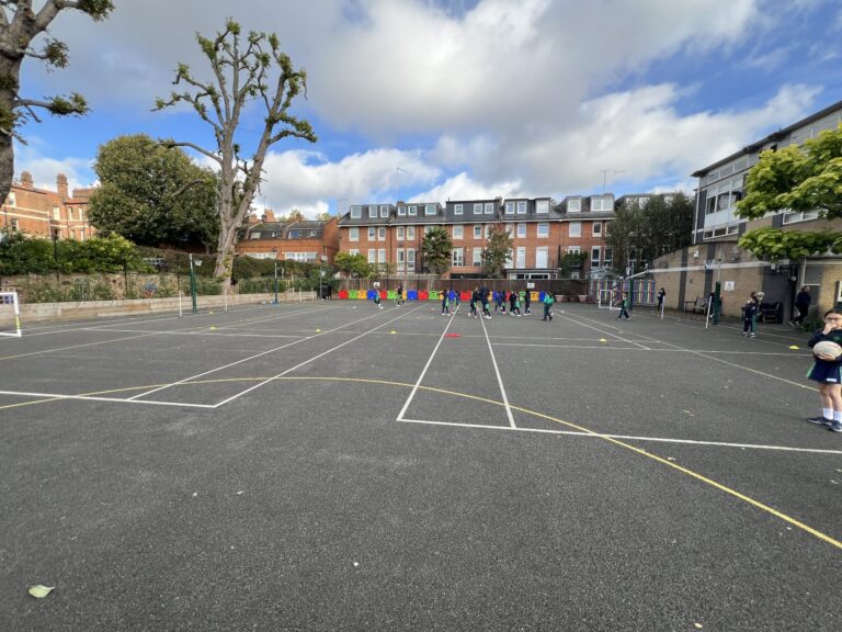 Children playing on a schoolyard with white court lines and colorful benches along a brick building backdrop under a cloudy sky, trees to the left.