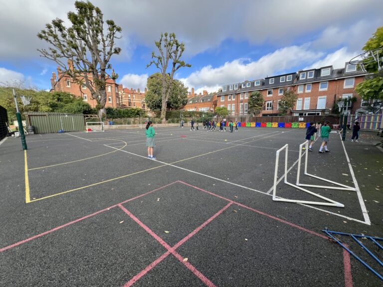 Children playing in a school playground with colorful barriers along the fence and chalk lines on the ground.