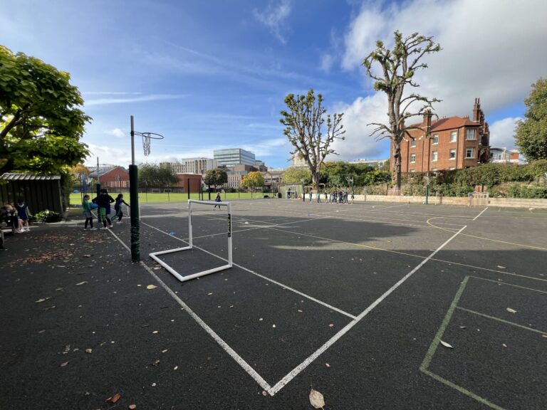 A school playground with a small white goal on a paved court, groups of children gathered on the left under green trees, blue sky and buildings in the background.