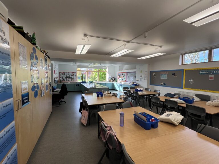 A bright classroom with long blue-topped tables and chairs arranged in groups, fruit-green cabinets on the left, and bulletin boards along the walls.