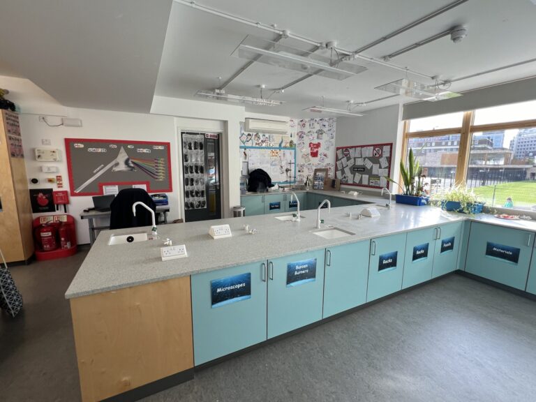 Bright science lab with turquoise cabinets along a long counter, labels for microscopes, rocks, fossils, and materials, and large windows to the right.