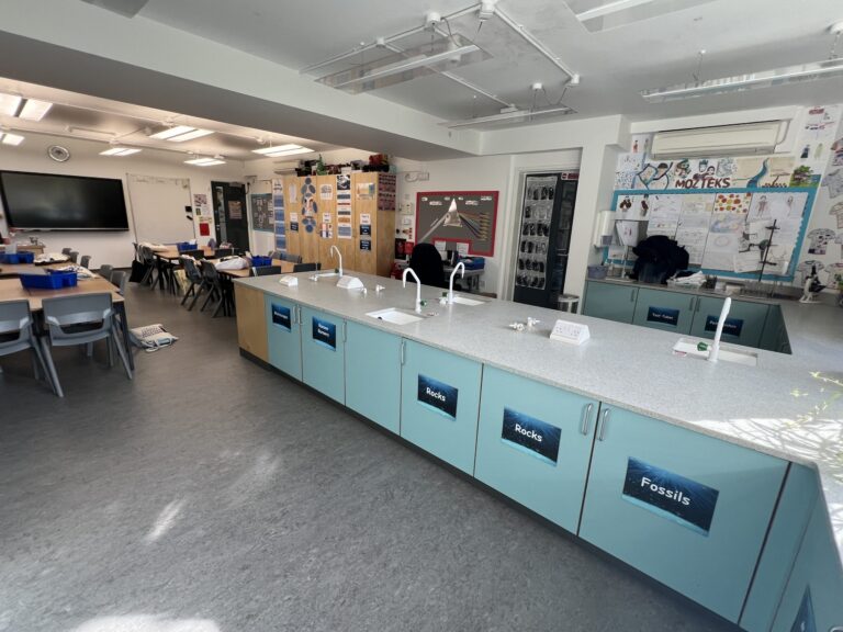Science classroom with blue lab cabinets along a counter, labeled Rocks and Fossils, and student tables on the left.