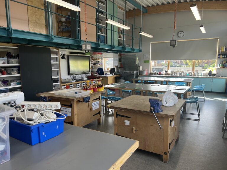 A spacious classroom/workshop with long workbenches, teal overhead storage, and shelves filled with supplies along the back wall.