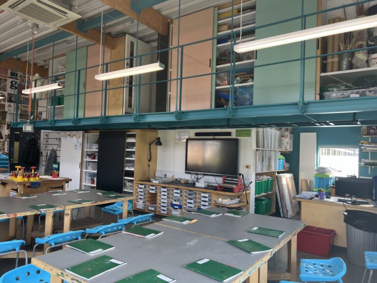 Art classroom/workshop with long tables, green mats, and blue plastic stools arranged for projects