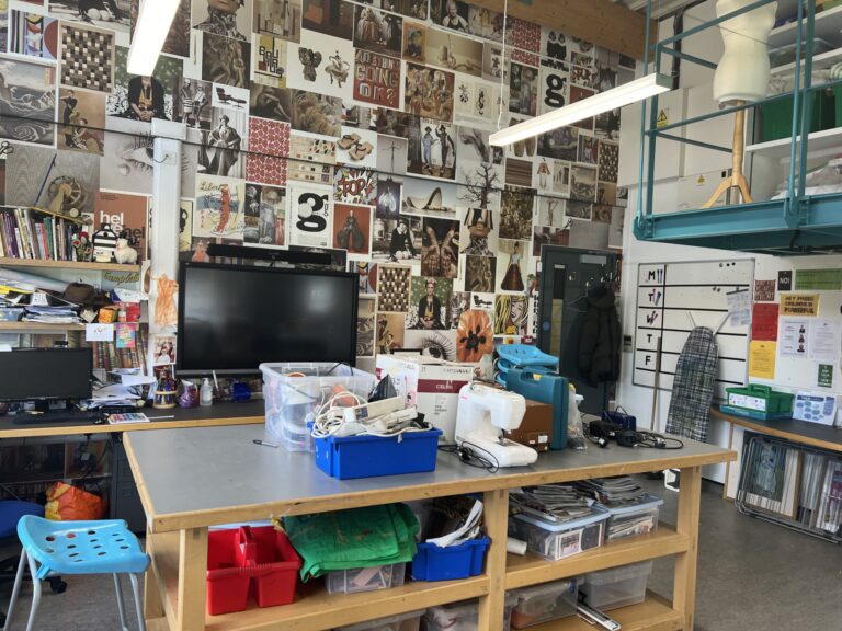 Cluttered workshop with desks, shelves, and a wall covered in posters and artwork collage behind the work area.