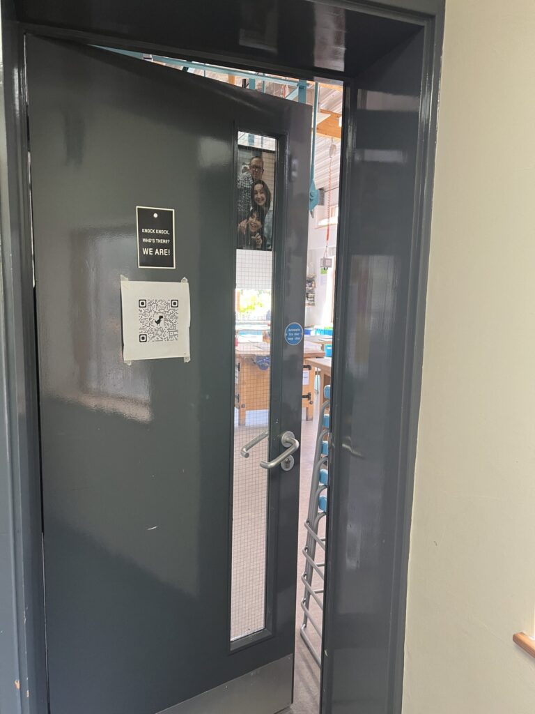 Dark gray door with a tall vertical window; three people peek in from the adjacent room, smiling at the camera.