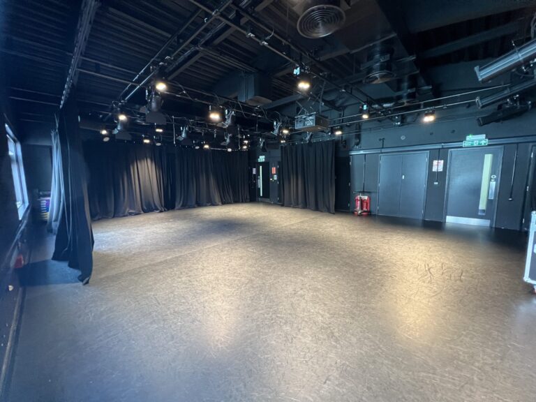 Backstage theater area with overhead lighting rig, spotlights, and black curtains along the walls; exit and fire safety equipment visible.
