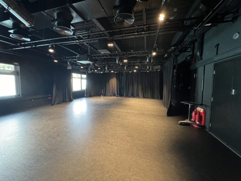 Interior of a black-box theater: empty stage with black curtains at the back and overhead lighting rig.