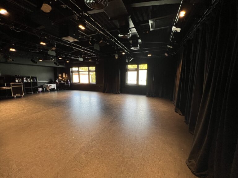 Empty rehearsal studio with black walls and stage curtain on the right, overhead lighting rig, and a few people at the back left near equipment. Windows at the far wall let in daylight.
