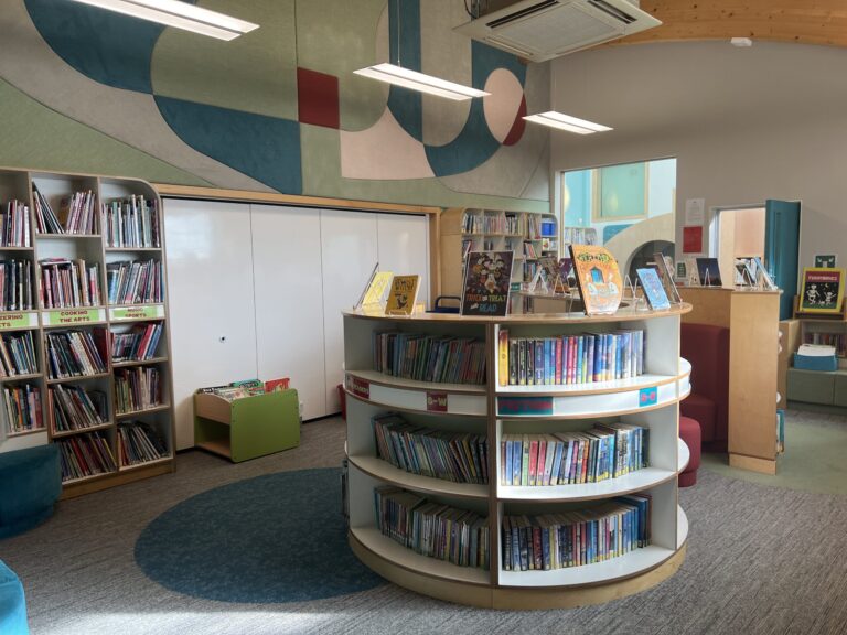 Curved white round book display filled with children's books in a colorful mural library walls and ceiling lights nearby.