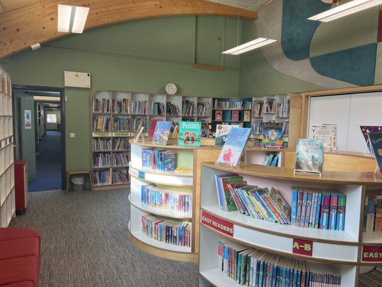 Children's library interior with curved white display shelves filled with colorful books and a green wall backdrop; a wooden ceiling and mural above create a playful atmosphere.