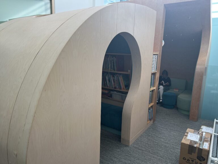 Curved wooden shelves form an arched library passage, with a person reading in the background near blue seating and glass walls.
