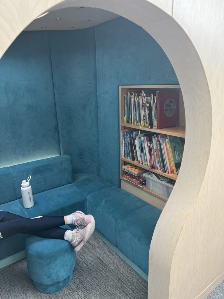 Blue curved booth seating forming a circular nook with a bookshelf at the side; water bottle on the seat and legs in sneakers nearby.