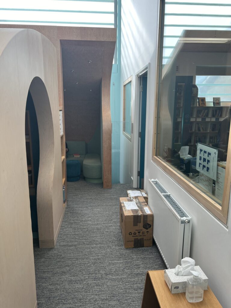 Library corridor with curved wooden partitions and a small seating area at the end, a cardboard box on the carpet nearby.
