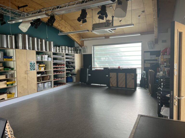 Industrial arts studio with tall metal storage canisters along cabinets, teal wall, and an overhead lighting rig, window at right.