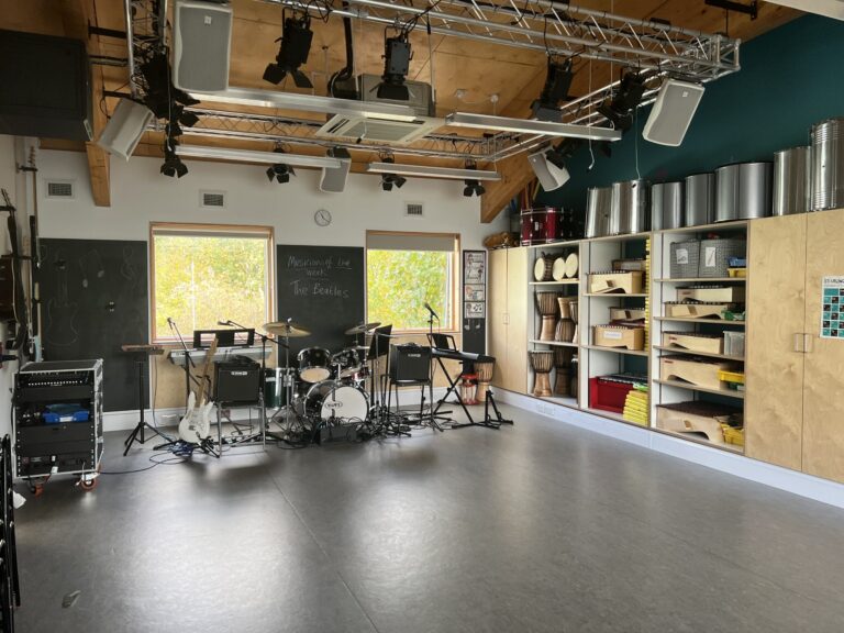 Music rehearsal room with a drum kit, guitars, amplifiers, and an overhead lighting rig.