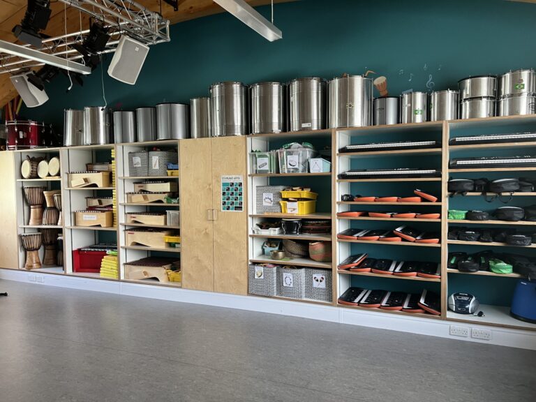 Music store interior with metal drums lined along shelves and various percussion and keyboard items on lower shelves under a teal wall.
