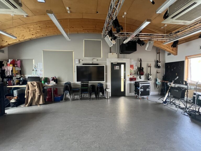 Wood‑ceiling music rehearsal room with a hanging lighting rig and several guitars on the wall, plus stacked chairs and equipment.