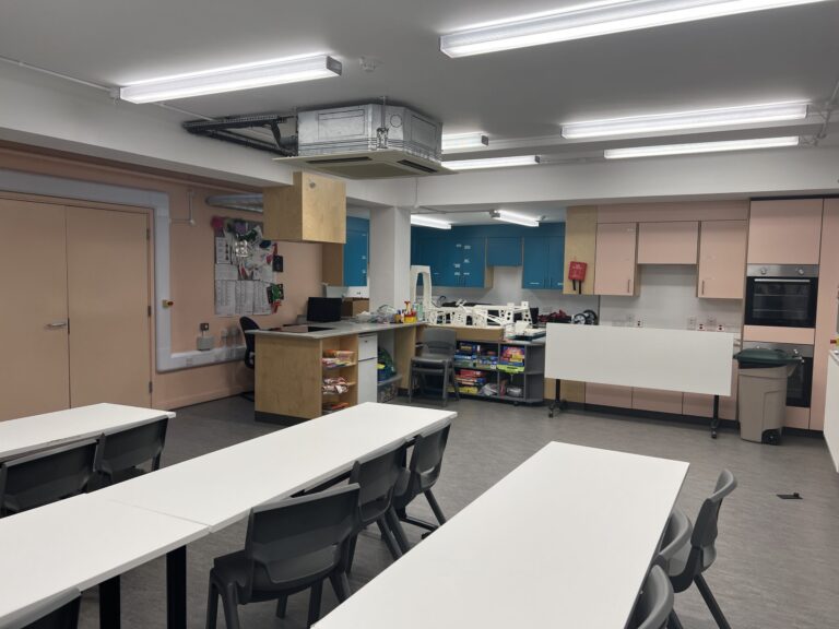 Open classroom or lab with long white tables, grey chairs, pink and blue cabinets, and exposed ceiling ducts.