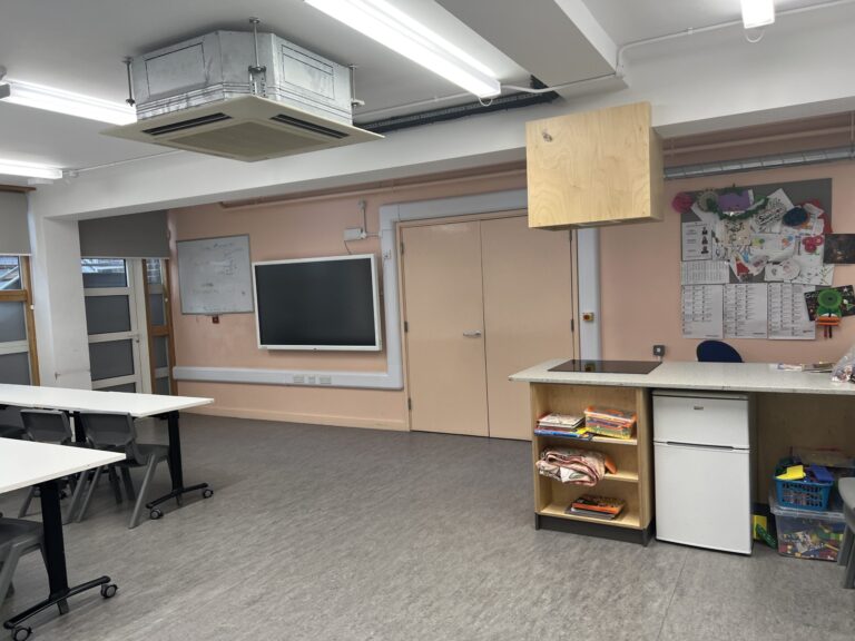 Classroom with a suspended HVAC unit, fluorescent lights, and pastel pink walls; tables and chairs arranged for a lesson.