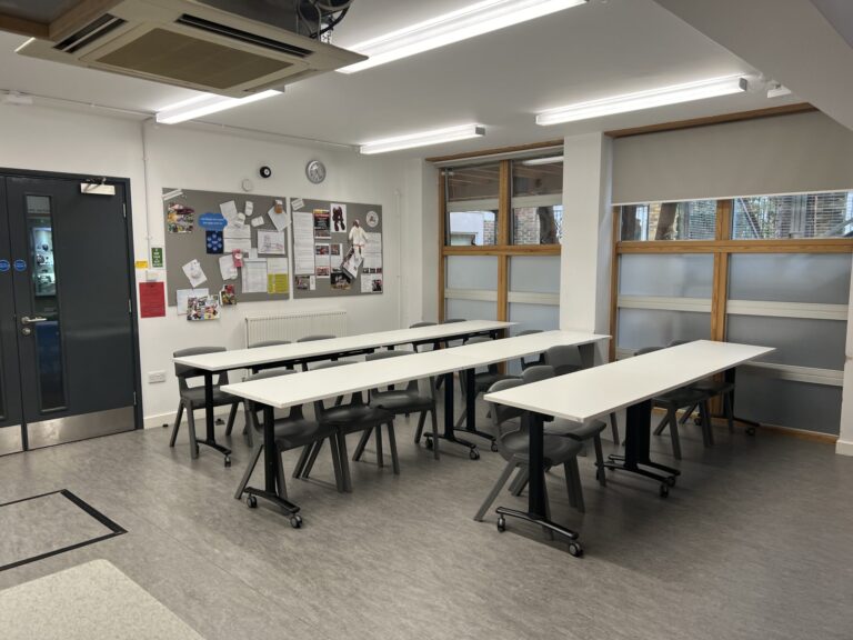 Bright classroom with long white tables on wheels, grey chairs, bulletin board, and large windows outside the room