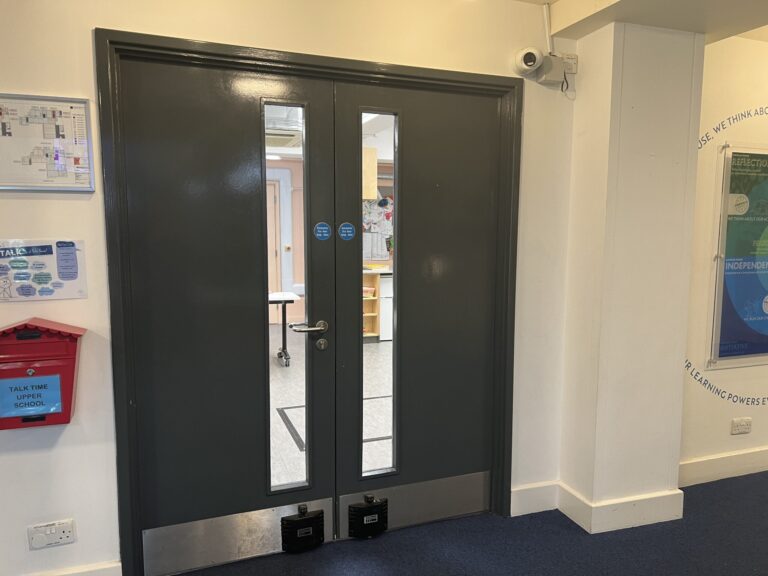 Double glass-panel doors in a hallway; view into a room with a table, a security camera above the door, and posters on the walls.