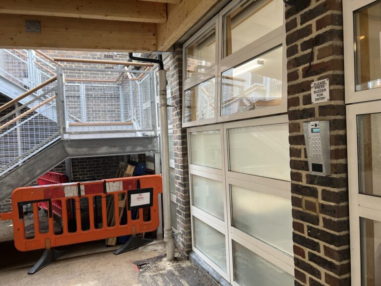 Construction area outside a glass-door entrance with orange safety barriers and metal stairs; a sign reads 'Please press buzzer for entry'.