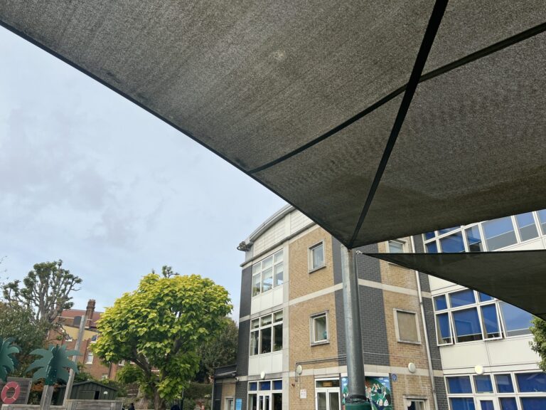 Shade sail canopy over a courtyard with a brick building and green trees visible below.