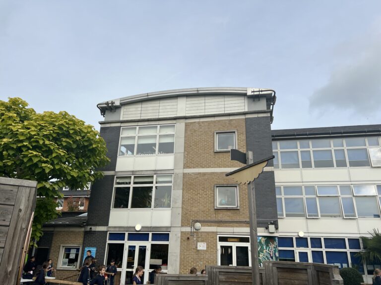 Front view of a brick school building with large windows; students in dark uniforms stand outside under a blue sky, with a leafy tree on the left.