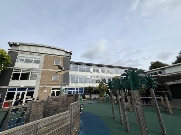Outdoor school playground with wooden climbing frames and blue-green safety flooring, a multi-storey brick-and-glass building in the background under a clear sky.