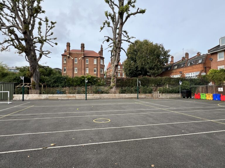 Empty schoolyard court with painted lines and circular markings, brick buildings and pruned trees in the background.