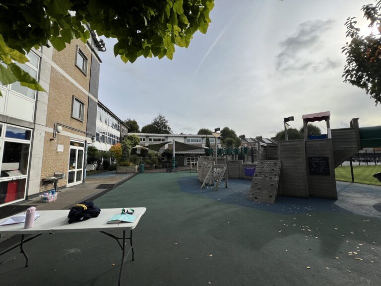 School courtyard with a climbing wooden structure, tables with papers, and a brick building on the left under a partly cloudy sky.