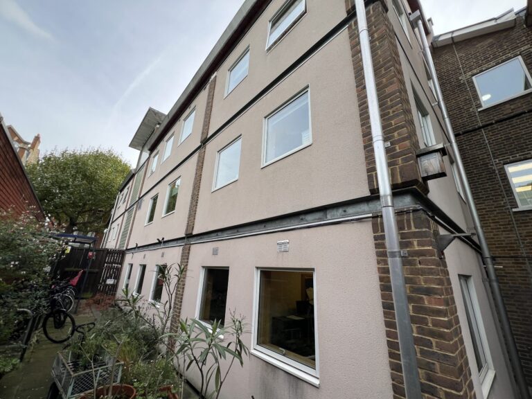 Beige multi-story apartment building with white-framed windows, brick corner accents, and metal downpipes; small garden and bicycles in front.