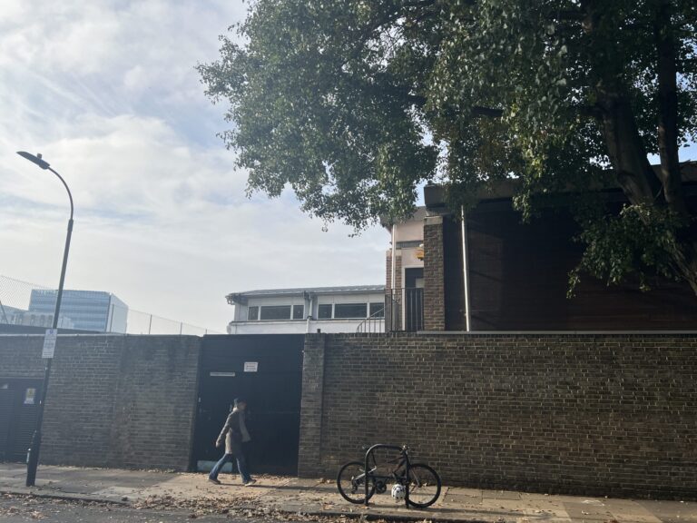 Person walking along a brick wall beside a parked bicycle on a city sidewalk beneath a large leafy tree and curved street lamp.