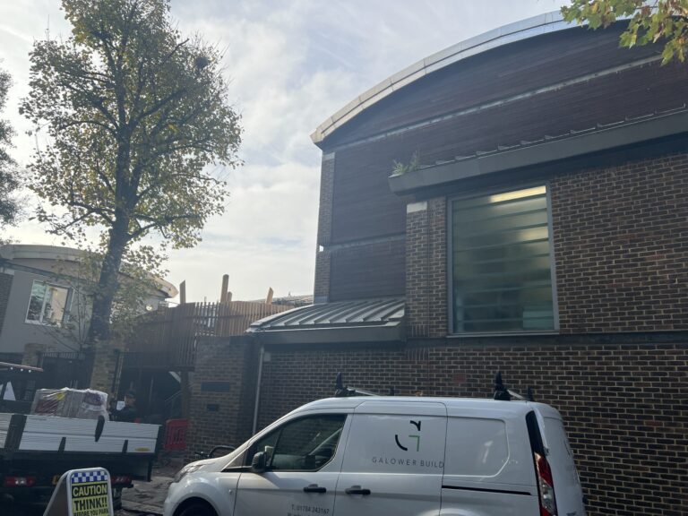 Brick building with a curved metal roof and large window, a tall tree to the left, and a white van marked GALOWER BUILD parked in front near construction barriers and a caution sign in the foreground.