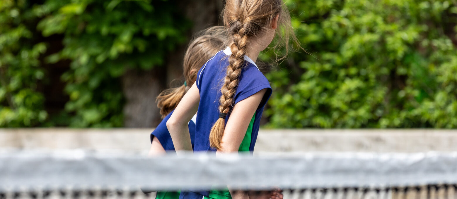 Girl in a blue and green sports uniform with a braided ponytail, walking outdoors beside a metal fence with green bushes behind.